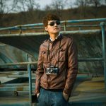 Stylish man wearing sunglasses and leather jacket holding a vintage camera in an outdoor setting.