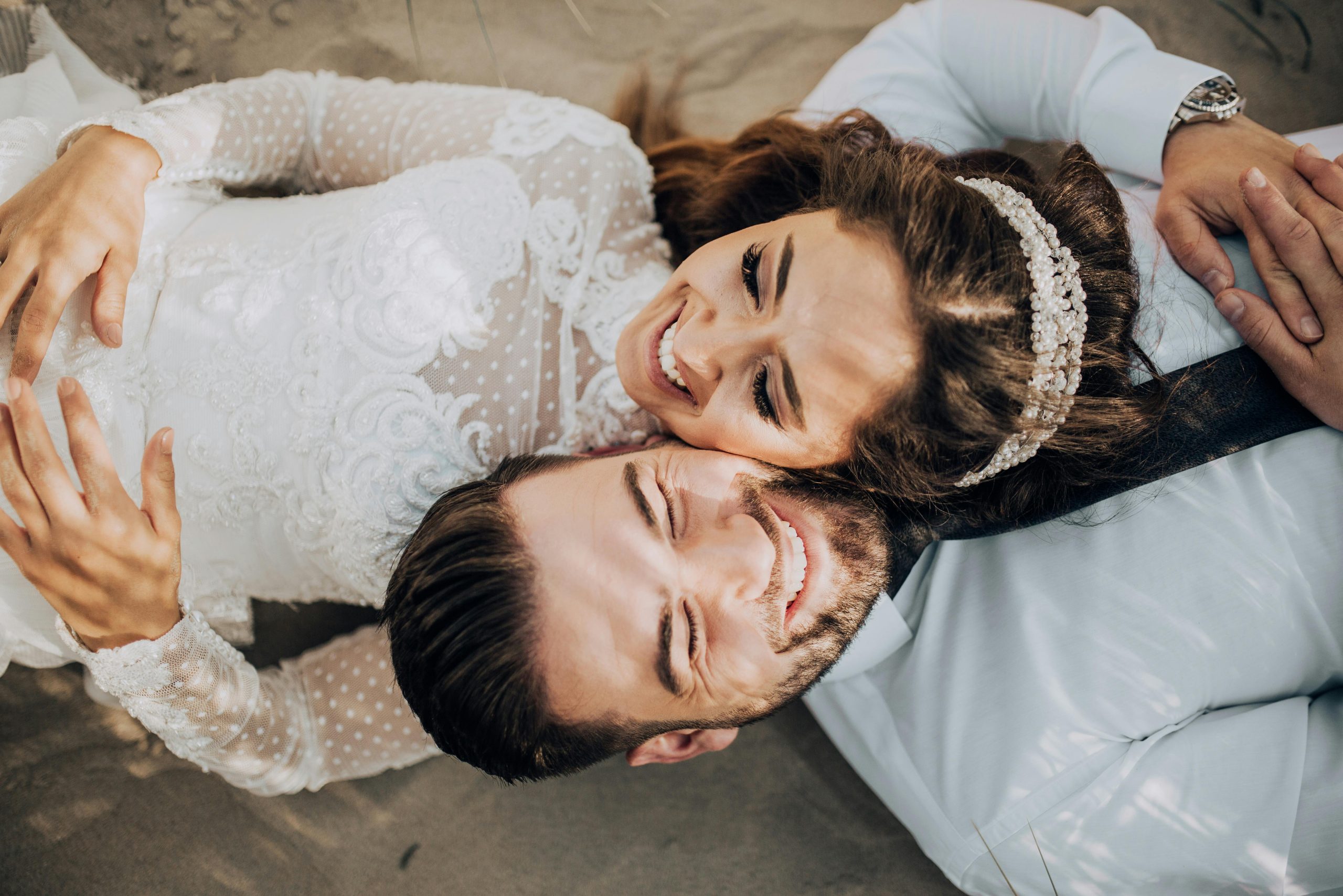 Happy bride and groom lying side by side on the beach in wedding attire. Capturing a joyful and intimate moment.