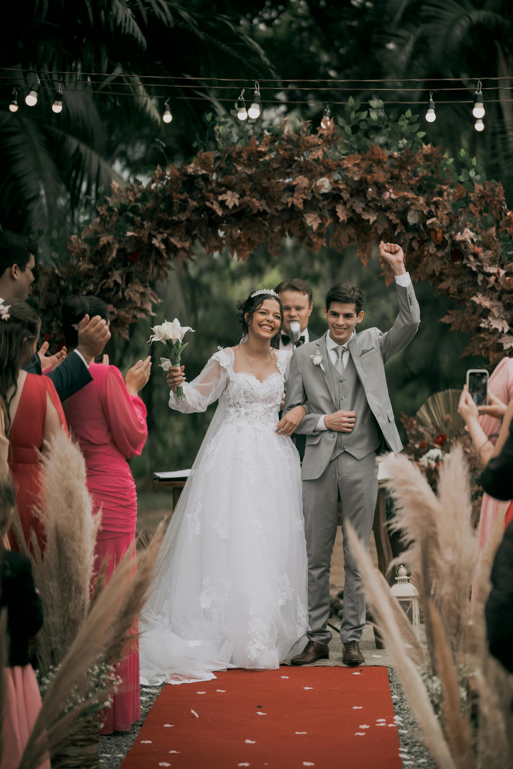Happy couple celebrates their wedding under a floral arch with loved ones cheering on a festive day.