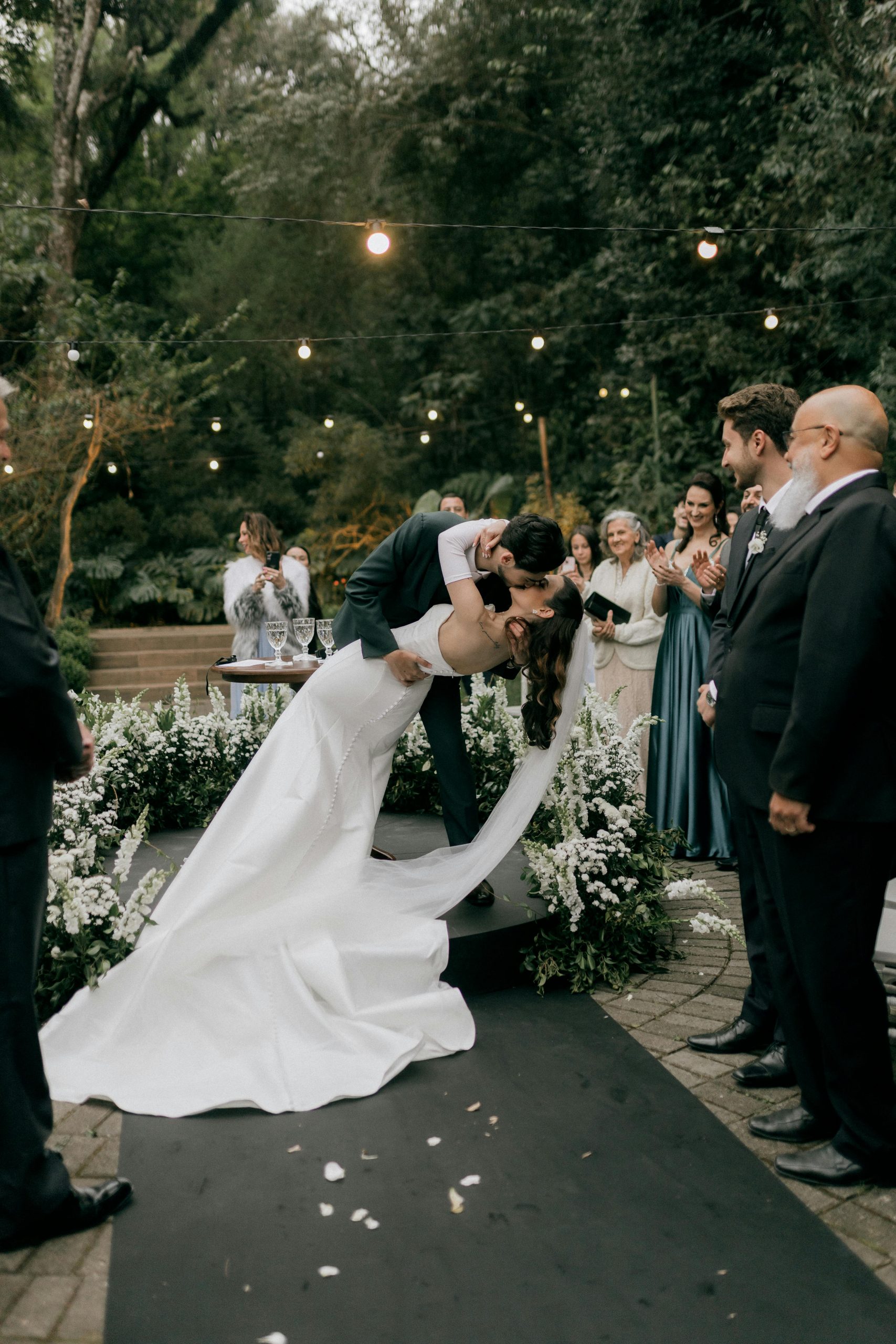 A couple's romantic kiss at an outdoor wedding ceremony surrounded by guests and flowers.